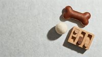 Dog toys flat lay on gray background: leather bone, wool felt ball, and wooden puzzle tray arranged with copy space for pet care content.