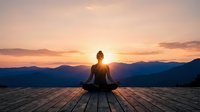 Woman meditating in lotus pose on wooden deck at sunrise with mountain landscape backdrop. Peaceful yoga practice for wellness and mindfulness concepts.