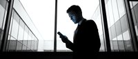 Professional businessman silhouette using smartphone in modern office building with glass windows. Corporate executive checking mobile device against urban skyline.