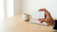 Young adult male with medium brown skin tone holding blank white business card at wooden desk with coffee mug and notebook, suitable for corporate branding and professional marketing materials.