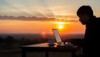 Young professional man working on laptop computer during golden hour sunset with warm orange sky and scenic landscape view from outdoor terrace or balcony.