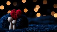 Romantic wooden figures embracing under red heart with bokeh lights creating magical atmosphere on dark blue textured background.