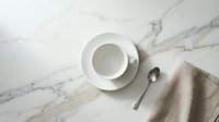 Empty white cup and saucer on marble surface with spoon and linen napkin, overhead view for café, restaurant, or tableware design projects.