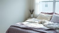 Unmade bed with layered bedding in neutral tones, white pillows, and dried flowers in vase by window with natural light for bedroom design and home decor.