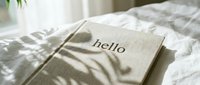 Linen-covered journal with hello lettering resting on white bedding in soft natural light with leaf shadows, cozy morning aesthetic.