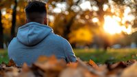 Young man in gray hoodie sitting alone in autumn park during golden hour sunset with fallen leaves around him creating peaceful contemplative mood for wellness content.