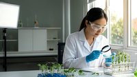 Asian female scientist in white lab coat examining plant samples with magnifying glass in modern laboratory for botanical research and analysis.