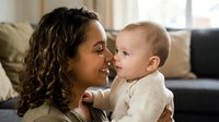 Young Hispanic mother with curly hair holding adorable baby in cozy living room. Tender maternal bonding moment showcasing love and connection between parent and child.
