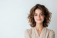Young Caucasian woman with wavy brown hair and natural makeup wearing beige wrap top smiling confidently against white background, positive emotional portrait.