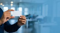 Businessman holding blank white business card in modern office environment with blurred background, suitable for corporate branding and professional networking materials.