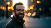 Happy middle-aged Caucasian man with beard and glasses smiling outdoors at dusk with warm bokeh lights creating atmospheric urban evening portrait.
