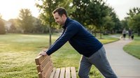 Active Caucasian man in sportswear stretching on a park bench outdoors during morning workout in green nature setting.