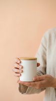 Person holding white ceramic container with wooden lid against neutral beige background for product mockup, branding, and lifestyle marketing purposes.