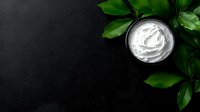 White moisturizing cream in glass jar with fresh green leaves on dark background, flat lay composition for cosmetic and skincare product photography.
