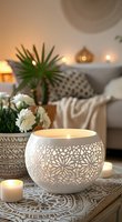Cozy home interior with glowing white ceramic candle holder featuring delicate floral cutout pattern, white chrysanthemums, and soft ambient lighting creating peaceful atmosphere.