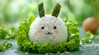 Cute bunny-shaped rice ball with nori ears nestled in fresh lettuce leaves, creative Asian food art against blurred green background. Fun kids meal presentation.