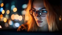 Young woman in glasses illuminated by warm bokeh lights at night, with dramatic moody lighting creating atmospheric portrait with glowing orange highlights.