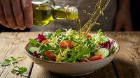 Olive oil being drizzled over fresh mixed green salad with tomatoes, cucumbers, and red leaves in white bowl on wooden table, healthy eating and food preparation concept.