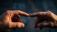 Two hands reaching toward each other with index fingers almost touching in warm golden light against dark blurred background symbolizing human connection and unity.