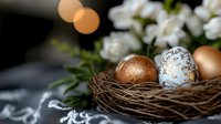 Decorative Easter eggs in natural nest with white spring flowers and bokeh lights. Golden and white painted eggs create festive holiday composition.