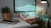 Hands holding glowing digital mesh wireframe hologram above desk workspace with blurred office background and potted plant, representing technology innovation.