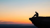 Silhouette of solitary person sitting on cliff edge against dramatic orange sunset sky and mountain landscape, contemplative peaceful moment in nature.
