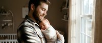 Young Caucasian father gently holds sleeping newborn baby by window in cozy nursery, capturing tender fatherhood and family bonding moment.