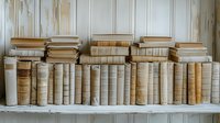 Vintage books with beige and cream bindings arranged on white wooden shelf against rustic paneled wall, creating nostalgic library atmosphere.