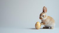 Adorable light brown bunny rabbit sitting next to decorated yellow Easter egg on neutral gray background. Minimalist holiday composition with copy space.