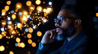 Young African American businessman in glasses contemplating against blurred golden bokeh lights background, profile view showing thoughtful expression and beard.