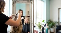 Professional hairstylist styling blonde woman's hair in modern salon with ornate mirror. Mother and daughter enjoying beauty treatment together.