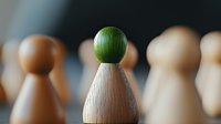 Wooden pawn figure with green top stands out from crowd of natural wood pieces on blurred background. Leadership and uniqueness concept for business presentations.
