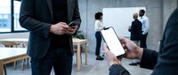 Professional businessman using smartphone in modern office meeting room with colleagues discussing project in background during corporate presentation.