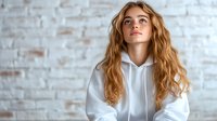 Young Caucasian woman with long wavy ginger hair wearing white hoodie sitting against brick wall background, looking up with dreamy expression. Natural lifestyle portrait.