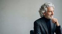 Mature man with gray wavy hair and facial hair, wearing dark blazer and black turtleneck, posing thoughtfully with hand near chin against neutral background for corporate and lifestyle use.