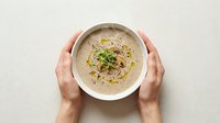 Hands holding bowl of creamy hummus topped with olive oil, paprika and fresh herbs, ready for serving or dipping.