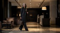Professional businesswoman walking through luxury hotel lobby with briefcase wearing elegant black suit and confident expression.