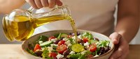 Man pouring golden olive oil over fresh vegetable salad with tomatoes, lettuce, and feta cheese for cooking and food preparation.