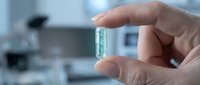 Hand holding transparent capsule pill in modern laboratory setting for pharmaceutical research and drug development.