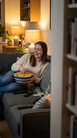 Happy young woman enjoying popcorn while watching TV on cozy living room sofa in warm evening light for entertainment and relaxation at home.