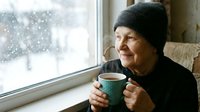 Senior woman in knit hat enjoying hot beverage by snowy window, cozy winter morning atmosphere, elderly lifestyle and seasonal comfort at home.