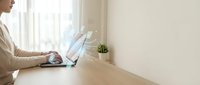 Professional woman working on laptop at wooden desk in bright modern office space with natural light from window creating productive workspace atmosphere.