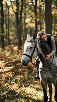 Woman with blonde hair and fair complexion riding gray horse on forest trail during autumn, wearing casual dark clothing and showing affection to animal in natural woodland setting.