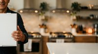 Man in dark blazer holding blank white sign in modern kitchen with holiday decorations and warm lighting for advertising or promotional messaging.