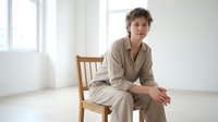 Young woman in beige linen suit sitting on wooden chair in bright minimalist room with natural light for business and lifestyle concepts.