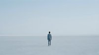 Solitary figure standing alone in vast white minimalist landscape with soft gray sky creating sense of isolation and contemplation for mental health concepts.