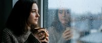 Woman with dark hair and fair complexion gazing pensively through rainy window, holding warm beverage, contemplative mood, urban setting, melancholic atmosphere for emotional storytelling.