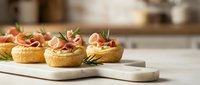 Gourmet puff pastry appetizers with prosciutto and herbs on white marble board in modern kitchen setting for catering and entertaining events.