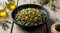 Fresh tabbouleh salad with parsley, tomatoes and bulgur in black bowl on rustic wooden table with olive oil bottles and herbs for healthy Mediterranean cuisine.