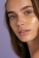 Young Caucasian woman with green eyes and freckles showing foundation swatches on cheek against purple background. Natural makeup look emphasizes glowing skin and beauty.